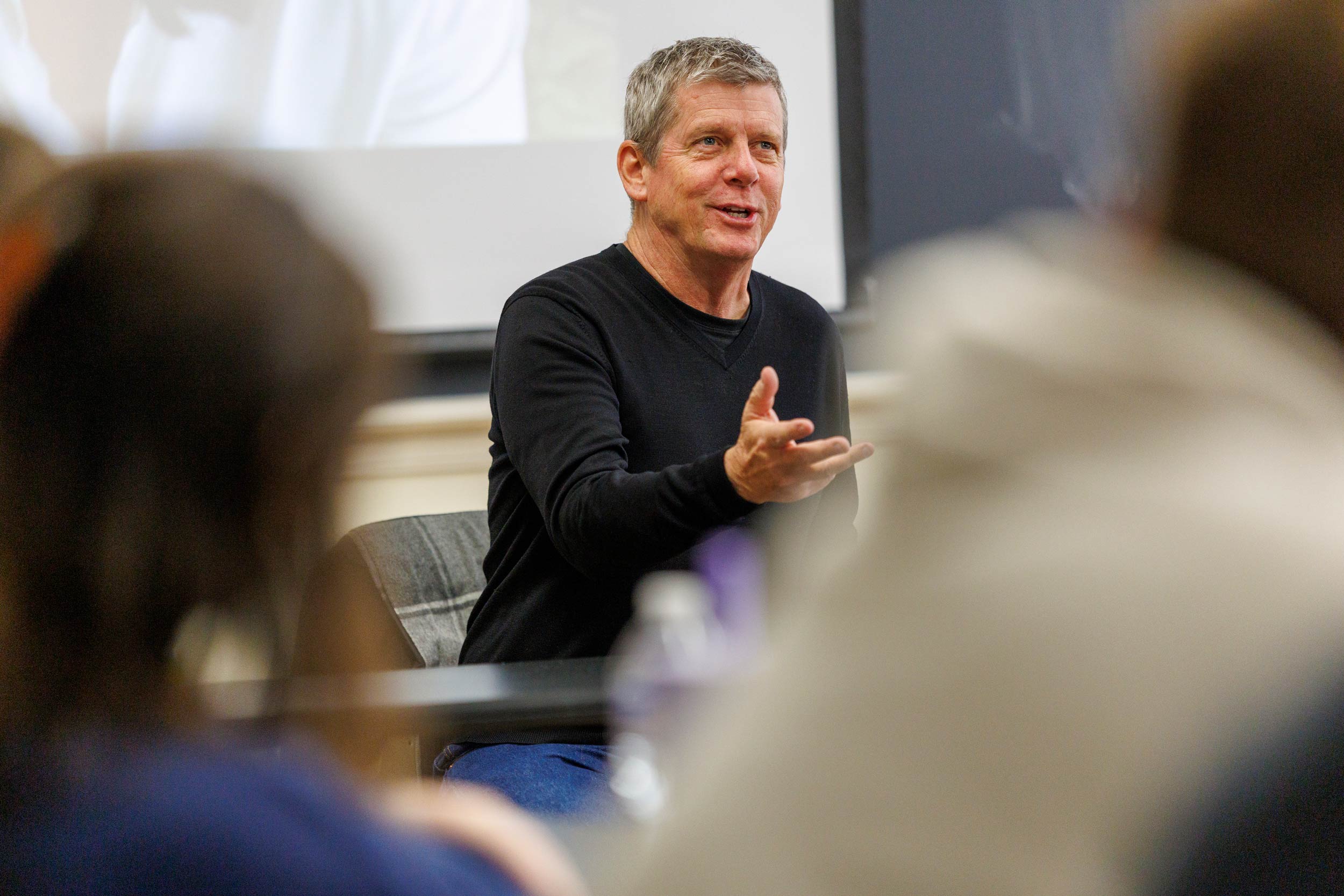 A middle-aged man in a black long-sleeve shirt sits at the front of a class and is gesturing forward towards the audience. There is a powerpoint being projected on a screen behind him.