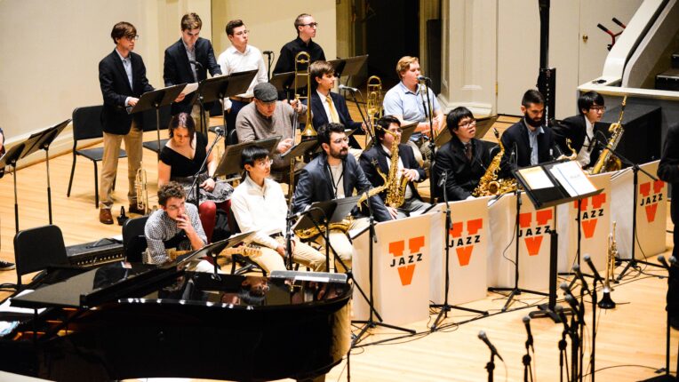 A group of jazz musicians stand and sit on a stage with various music stands and signs for UVA Jazz.