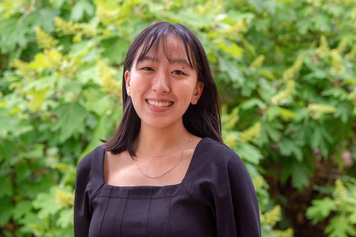 Photo of Jacquelyn Kim, brown hair with bangs and wearing a black top, stands in front of green foliage.