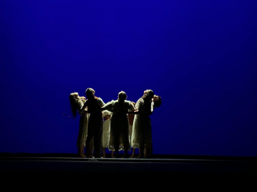A dark blue background behind a vast stage, with a small group of dancers in the center, all holding hands in a circle and looking up at the ceiling.