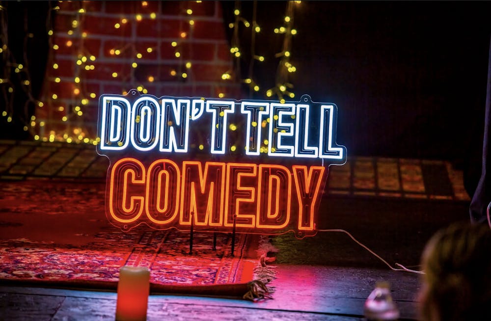 A stage with a red rug and a small neon sign that says "Don't Tell Comedy" in white and red letters.