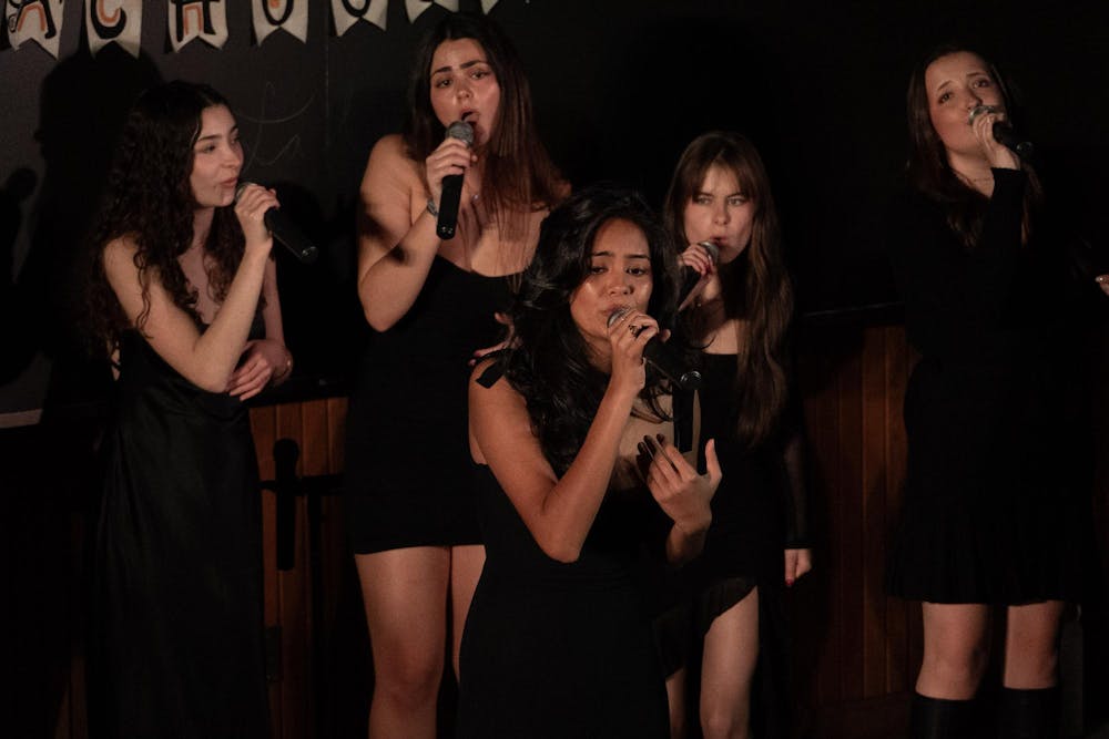 Five young women are shown singing into microphones on stage, standing close together, all wearing black clothing. 