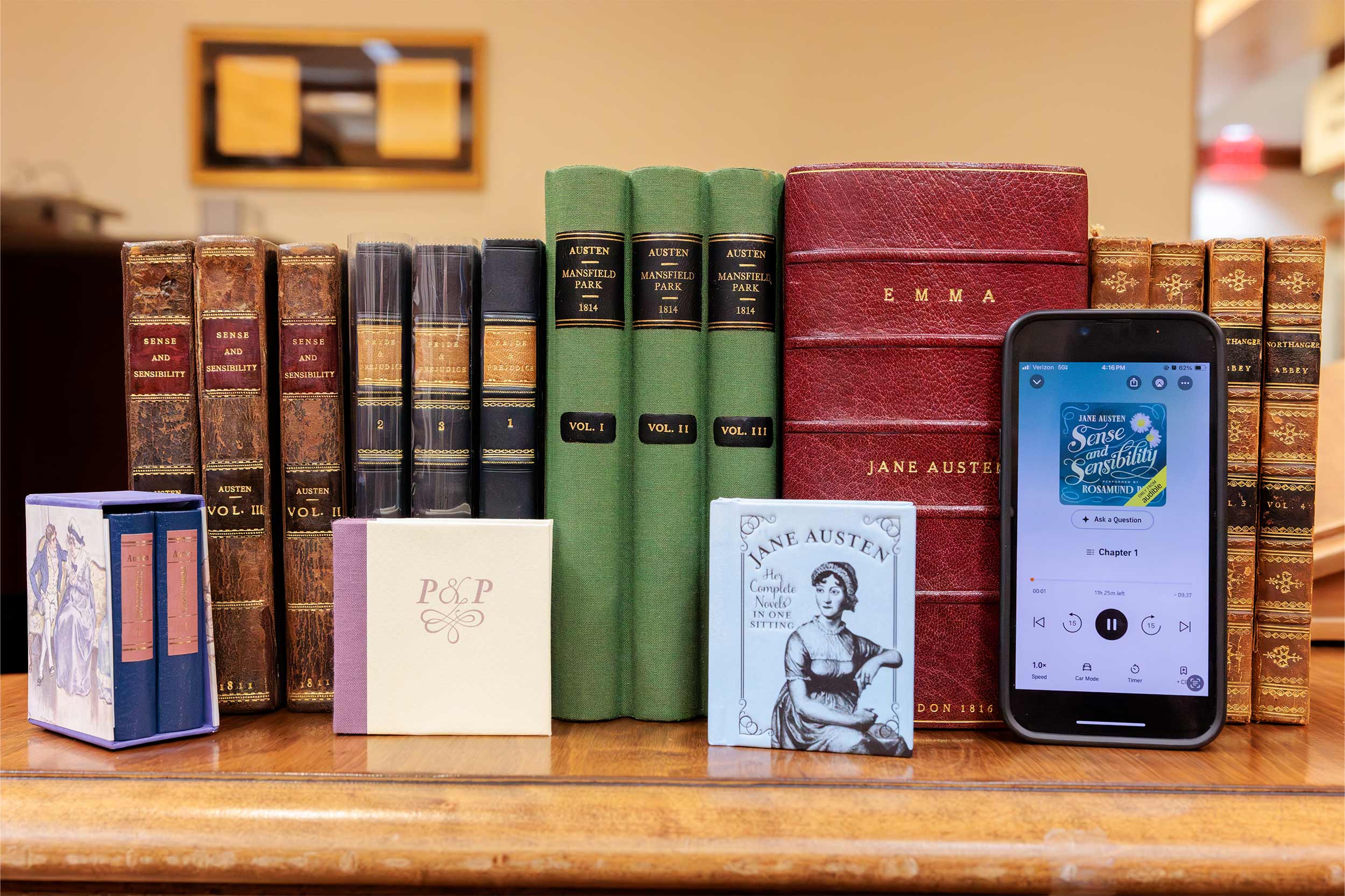 A line up of books on a table with their spines facing the camera, and a phone with a Jane Austen novel on the screen is leaning against the books.