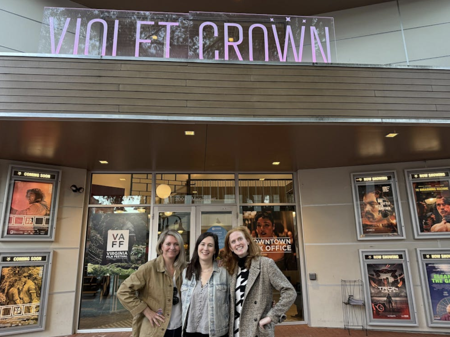 Three women stand in front of a movie theatre with a large sign, "Violet Crown" above them. The outside wall is filled with various movie posters.