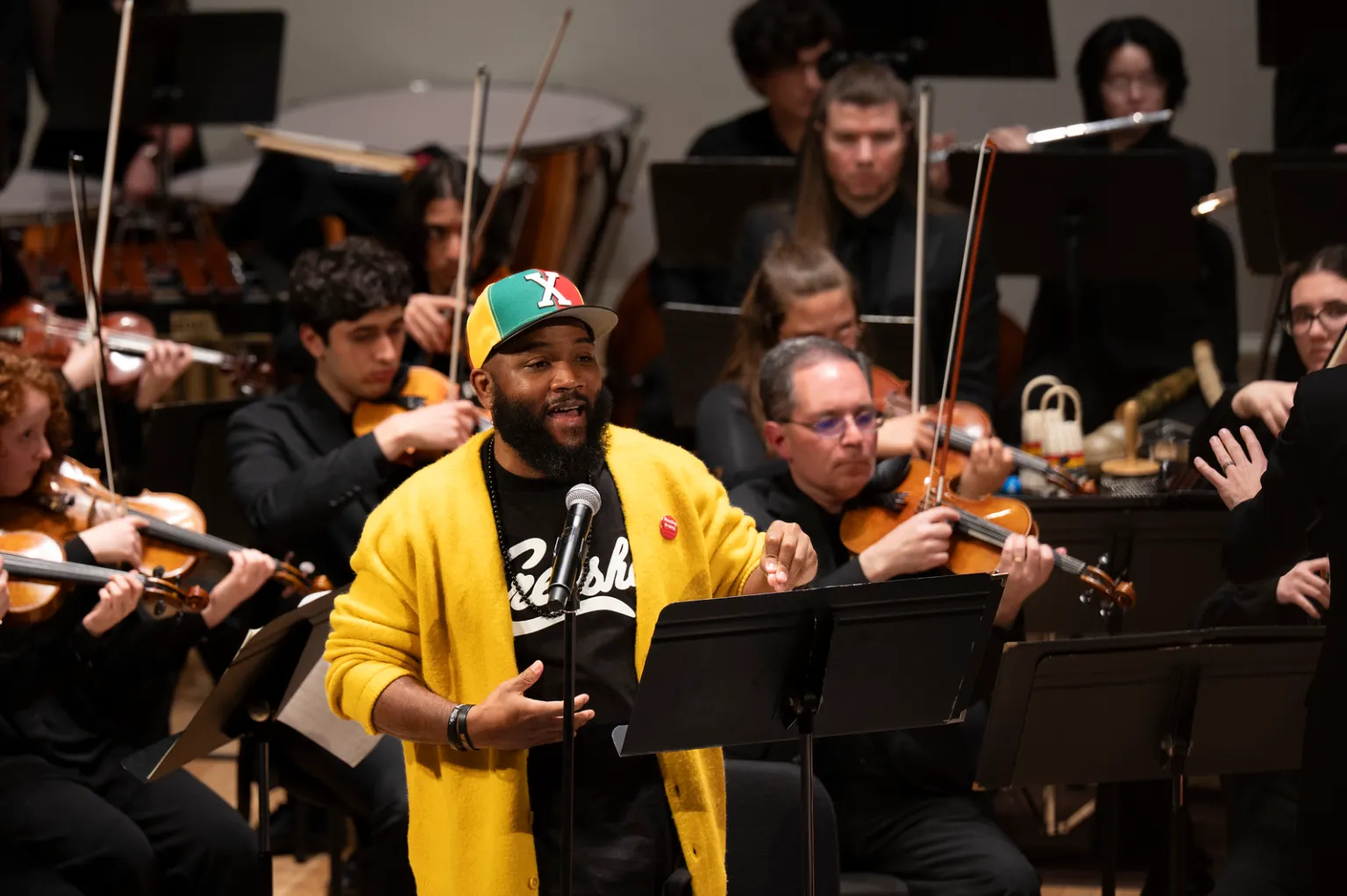 A man in a bright yellow sweater stands at a podium with a mic and raps to an audience, with a seated orchestra playing behind him. 