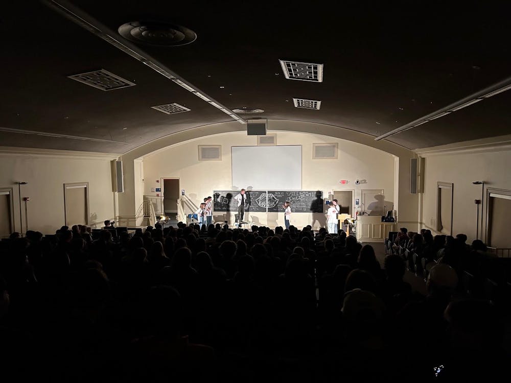 A dark auditorium with lights on at the front of the room where a group of performers stand while interacting with the chalkboard. 
