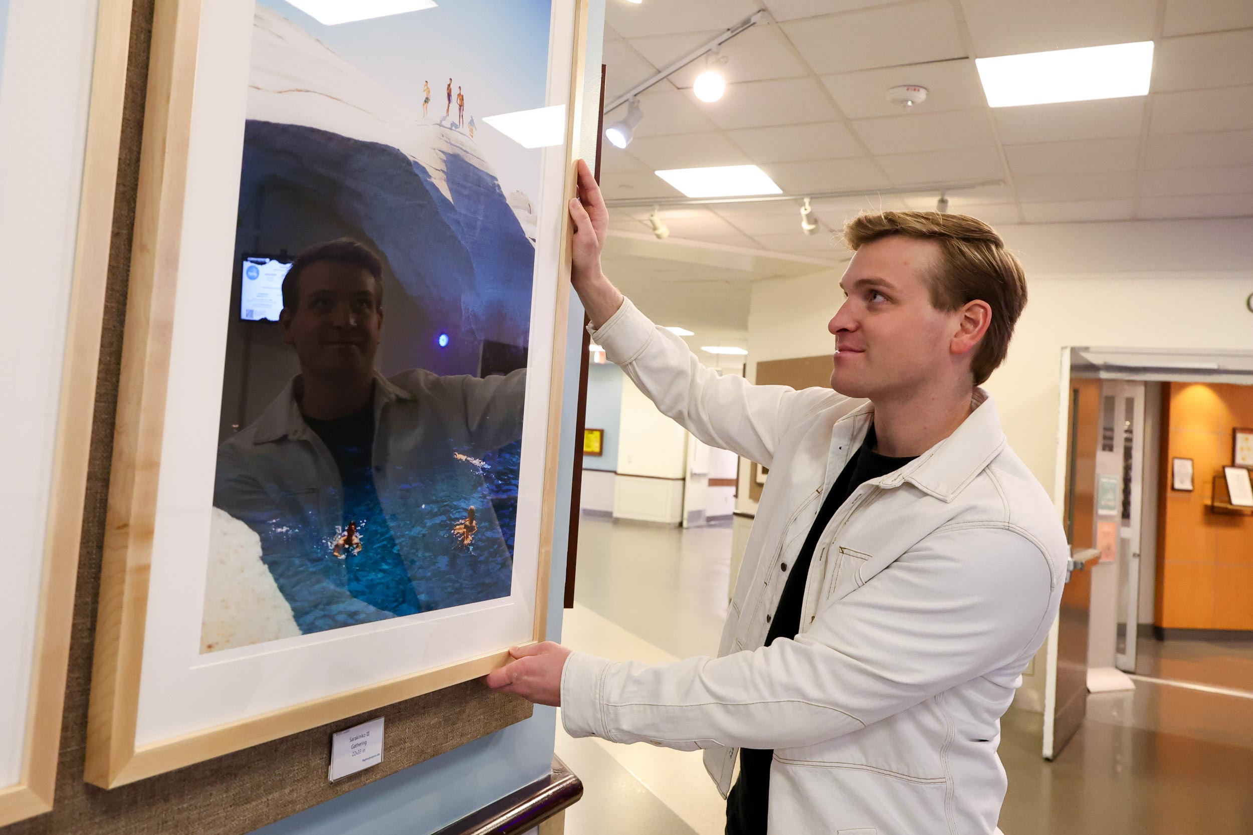 A blonde man in a white jacket adjusts a large framed photograph in a hallway, admiring the picture. 