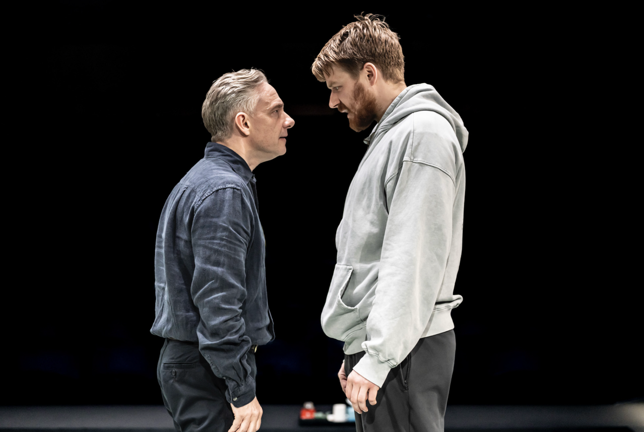 Two men stand closely in front of a black background and stare directly into eachothers' eyes.