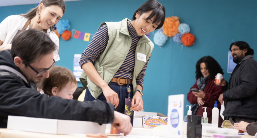 A bright blue room with tables and six visitors engaging in crafting activities. There is a young woman with a name tag helping a younger child.