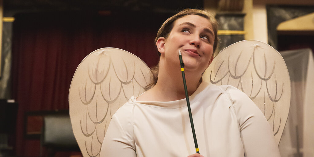 A blonde woman with a white shirt and white angel wings looks quizzically to the side, and ponders with a thin pointing stick tapping on her chin.