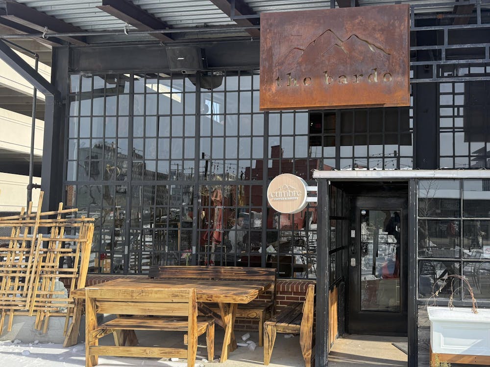 The front of an industrial urban building with a wall made of windows. A rusted sign hangs above the door and wooden tables and chairs are placed in the front.