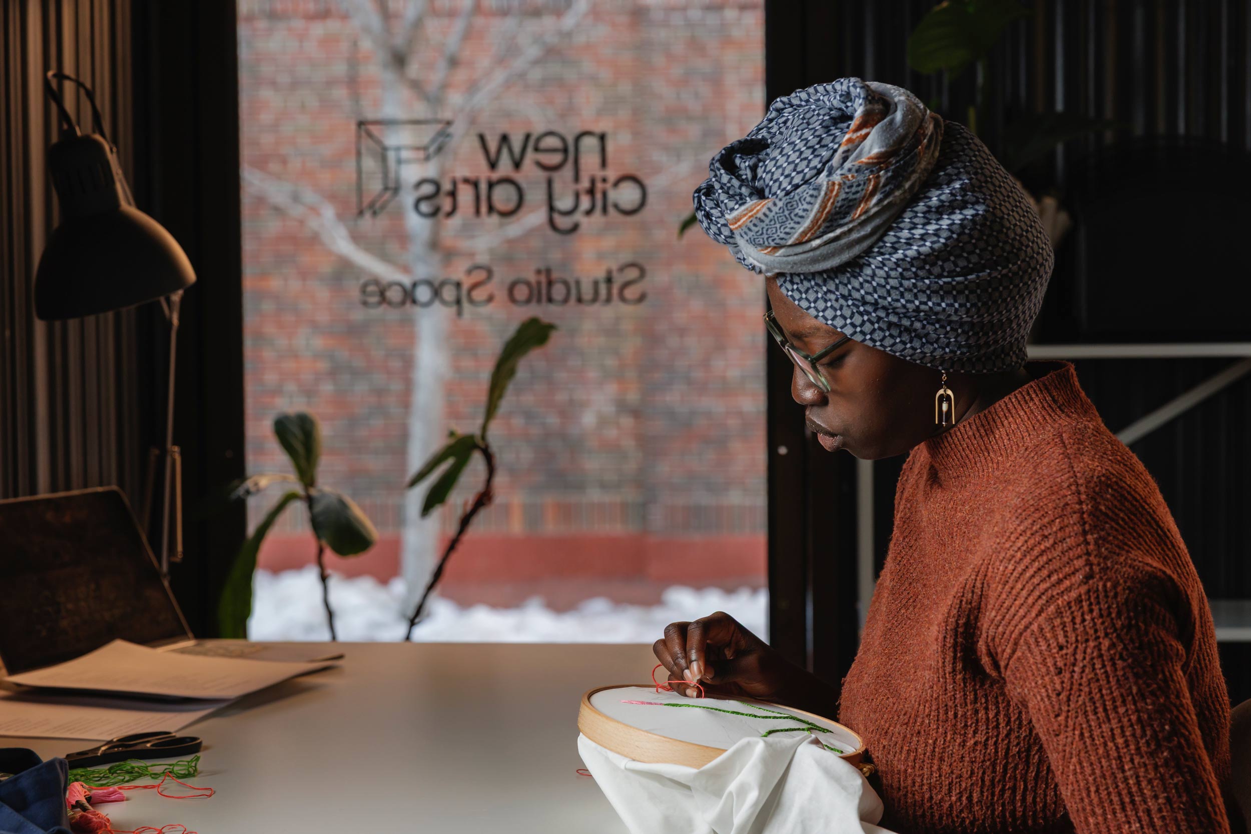 A young woman in an orange sweater and blue hair wrap sits at a table while practicing needlepoint.