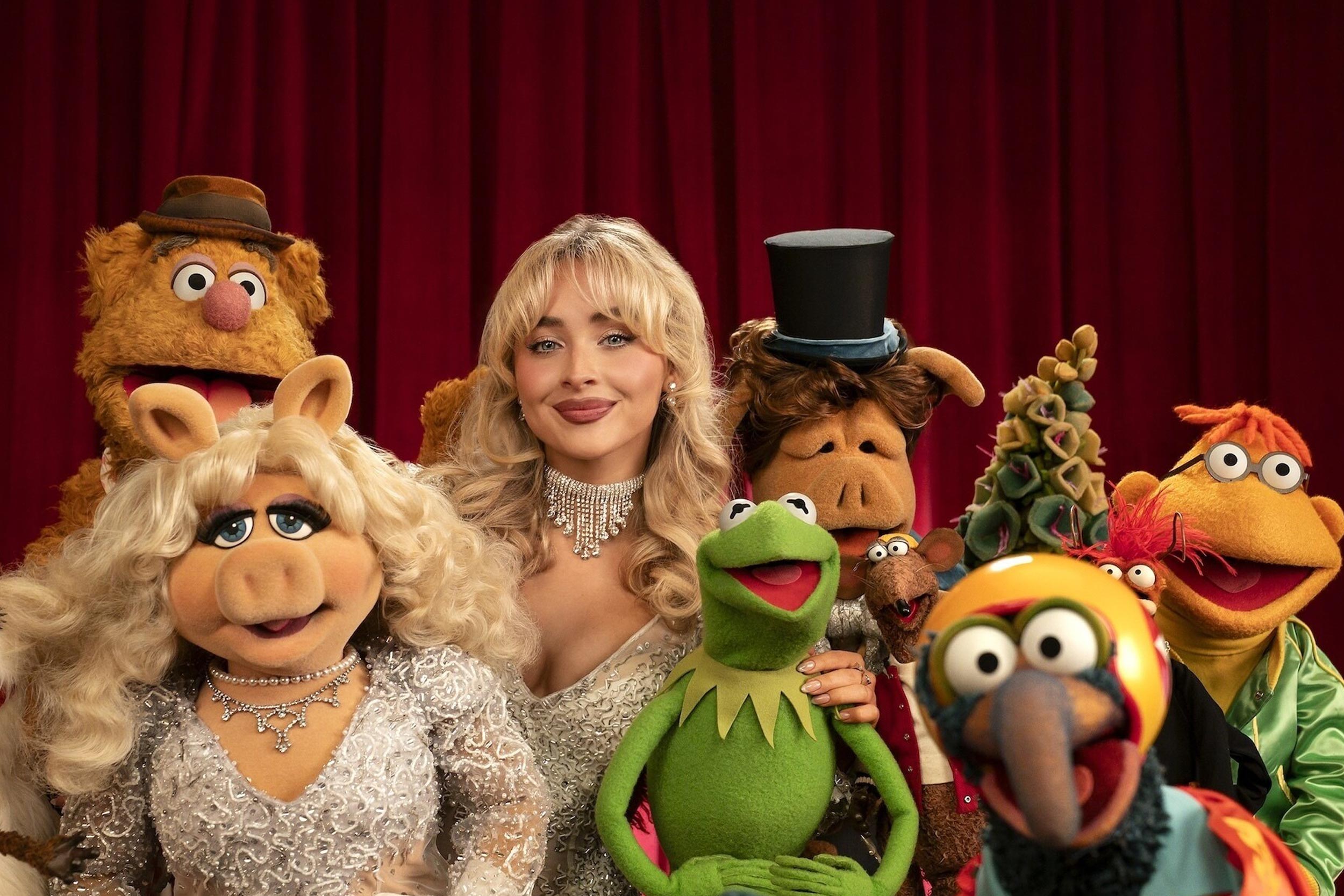 A red stage curtain with the main characters from the Muppets standing in front, smiling at the camera. Sabrina Carpenter is posed in the middle of the group.
