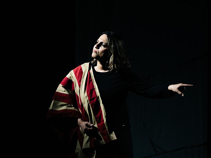 A woman stands on a completely dark stage with a spotlight illuminating her face and half of her body. She has a red and white striped fabric draped on her shoulder, and she is forcefully pointing to the side with a serious expression on her face.
