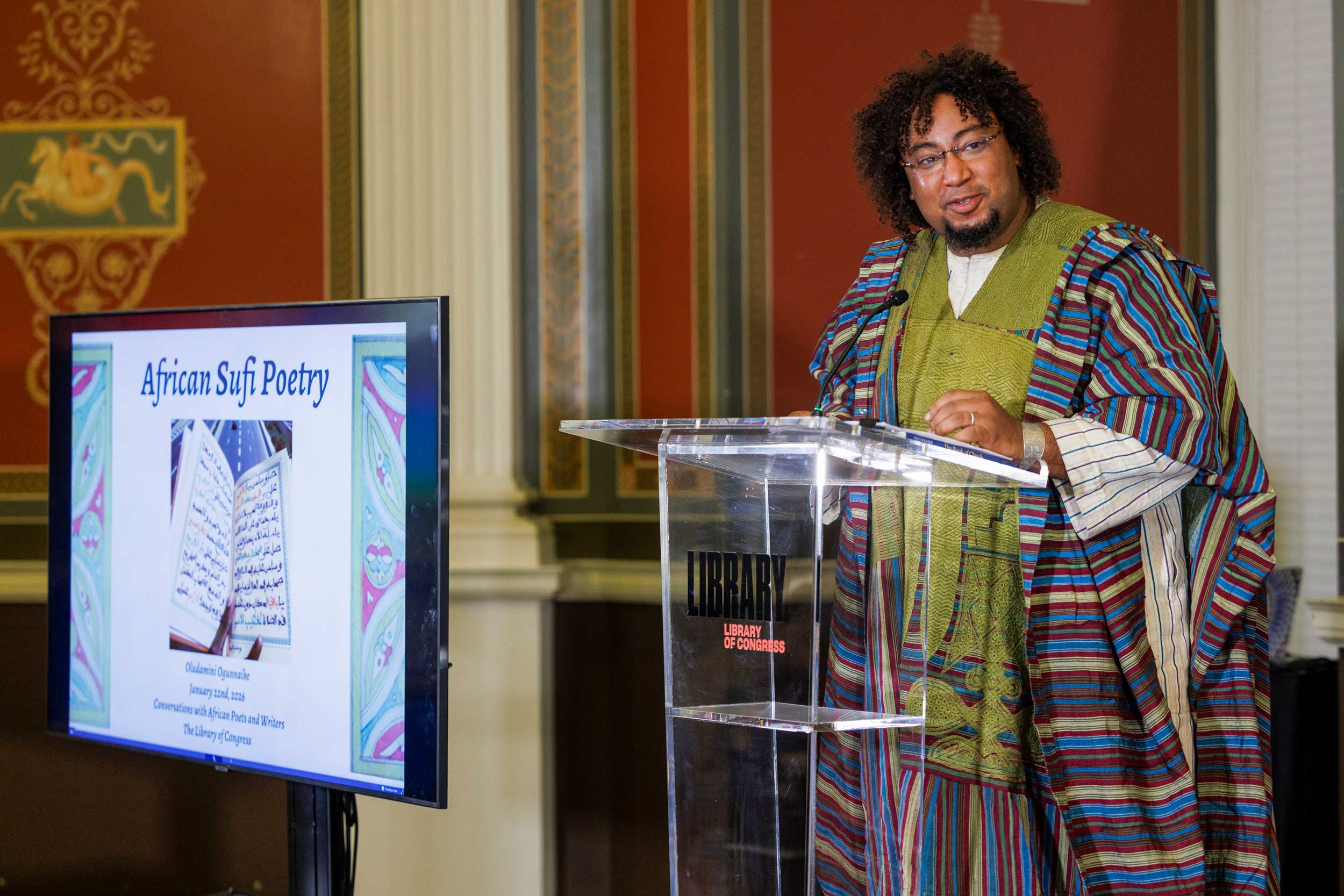 A man stands at a glass podium. He is wearing a colorful robe with patterned fabrics. As he is speaking, a screen next to him displays the title, "African Sufi Poetry" with a photo of an open book with writings inside.
