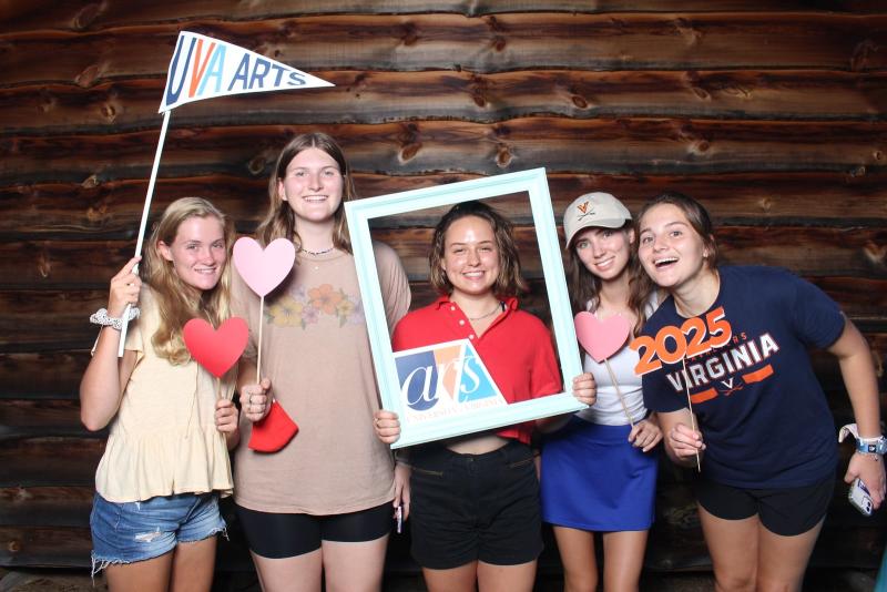 A group of five students stand in front of a log background in a photo booth, holding various signs in their hands. 