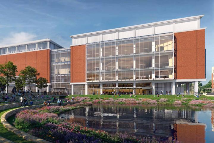 A large brick building with huge windows down the entire wall, with a small body of water in front of the building and outdoor seating within the nature.